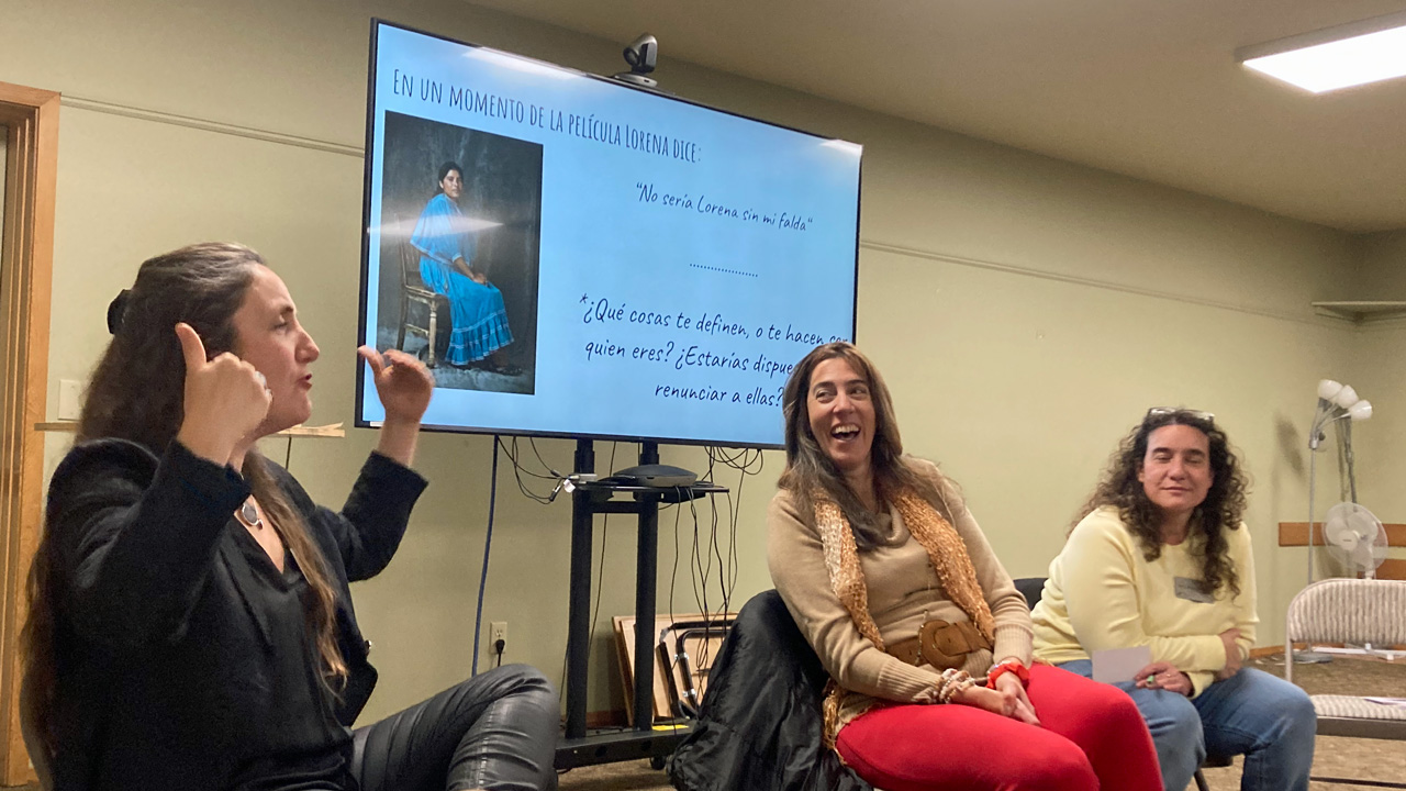 A photo of three women engaged in animated conversation, seated, with a projector screen behind them