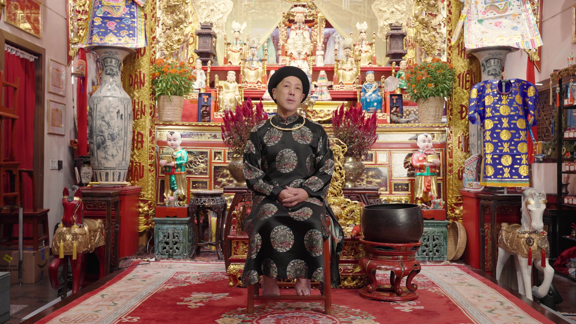 Allen Đức Lương, wearing a gold-embroidered robe, sits amid statues, flowers, and ornate furniture