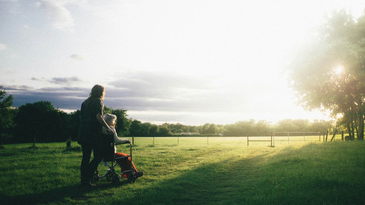 A photo of two women in a field, facing toward the sun, one pushing the other's wheelchair