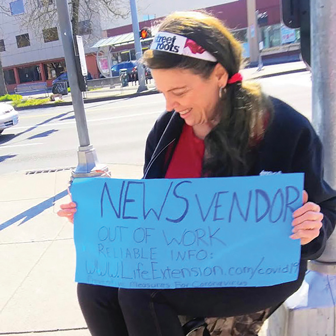 A woman sits on a street corner holding a sign reading, "News vendor. Out of work. Reliable info: www.lifeextension.com/covid19. Protective measures for Coronavirus."