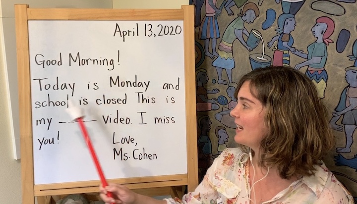 A woman gestures with a pointer toward a board that reads, "Good morning! Today is Monday and school is closed. This is my _____ video. I miss you! Love, Ms. Cohen"