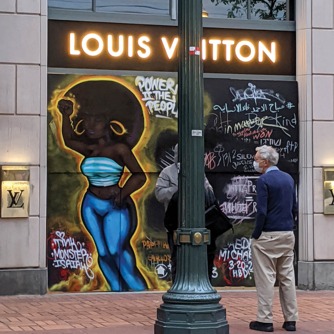 A photo of a street in downtown Portland. The Louis Vuitton store is boarded up, and a mural honoring Breonna Taylor is painted on the boards. An older white man wearing a mask looks at the mural.