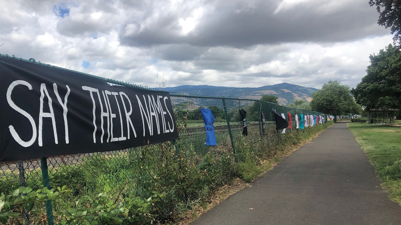 Before a southern Oregon landscape, a sign reading "Say Their Names" hangs on a fence along with T shirts bearing the names of Black men and women murdered by police.