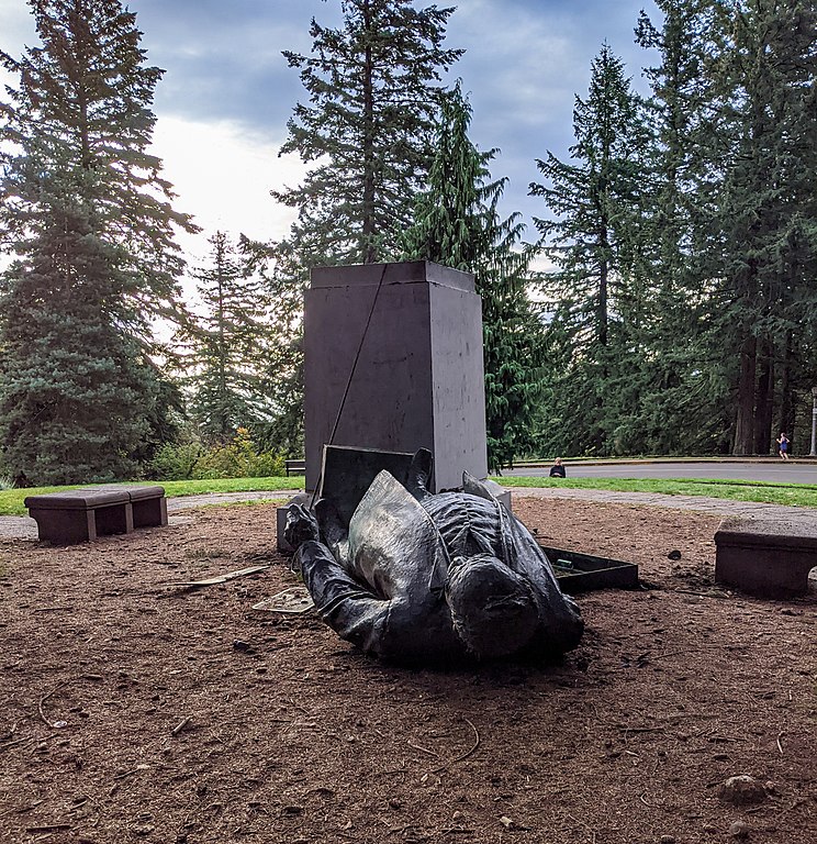 A photograph of the toppled statue of Harvey Scott, lying on the ground next to an empty pedestal