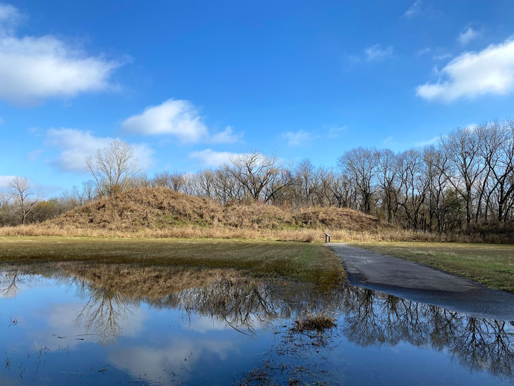 A photograph of an earthen mound with three humps seen from across a pond